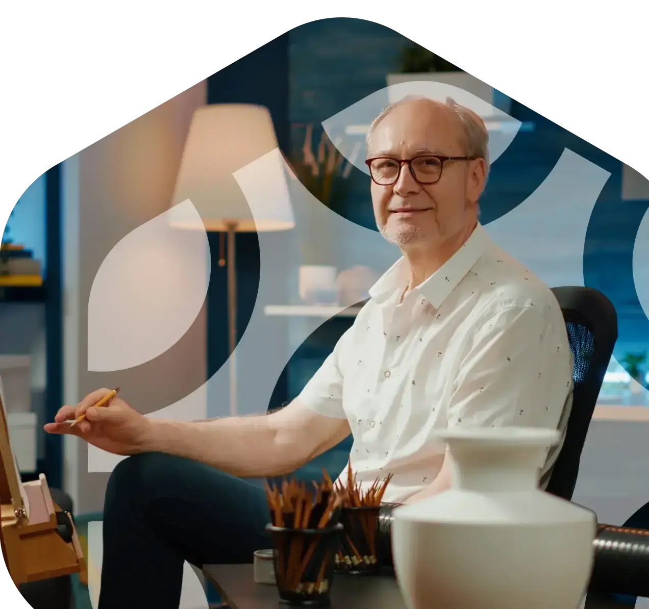 An image of a smiling older man seated, painting on an easel, framed in a watermarked version of the hexagonal logo shape.